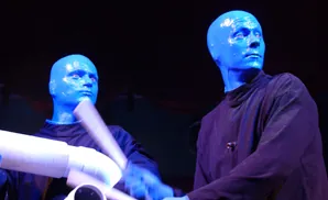 Blue Man Group performers, one with arms blurred in motion playing on white tubes.