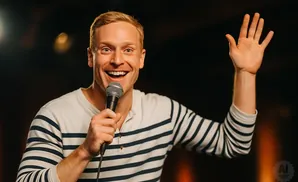 A man with blonde hair and a striped shirt smiles and holds a microphone, raising his hand.