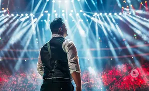 Man in vest and white shirt facing away from camera on stage, under bright blue and white spotlights, with a cheering crowd below.