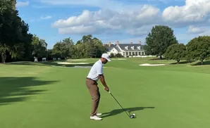 Golfer tees off on a sunny day with a clubhouse in the background.