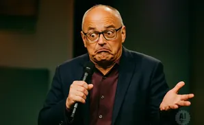 A man in a suit and glasses makes a surprised face while holding a microphone and gesturing with his hand.