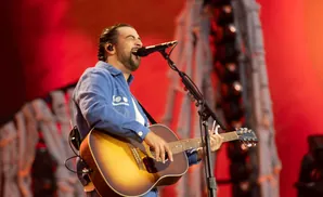 A man with a beard sings into a microphone while playing an acoustic guitar on stage.