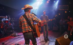 Musicians on a dimly lit stage with bright spotlights, one playing guitar and singing into a microphone, another playing guitar.