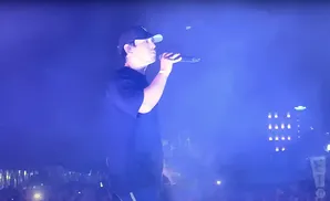 A man in a black t-shirt and baseball cap sings into a microphone on a brightly lit stage.