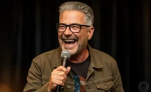 A man with graying hair, glasses, and a beard laughs into a microphone, wearing a green jacket over a t-shirt with an American flag design.