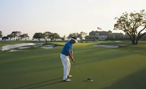 Golfer on a green prepares to hit a ball, with a large house and trees in the background.