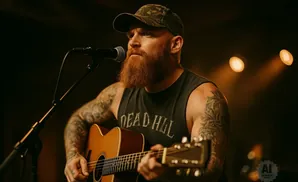 A man with a beard and tattoos plays an acoustic guitar while singing into a microphone on stage.
