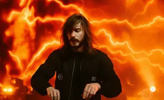 A man with long brown hair and a beard stands before an orange, lightning-filled background, wearing a black jacket.