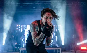 A male singer with tattoos on his arms and neck sings passionately into a microphone on a smoky stage with bright lights.