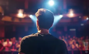 Man facing away from camera, on stage with spotlight, audience blurred in background.