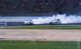 Two white drift cars with blue flames are creating large smoke plumes on a race track, with spectators in the background.