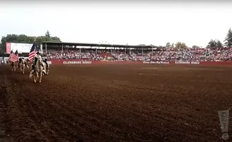 Rodeo parade with riders carrying American flags in front of stadium seating.