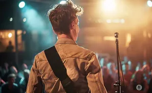 A man with messy hair plays a guitar on stage, facing away from the camera, with a microphone in front of him.