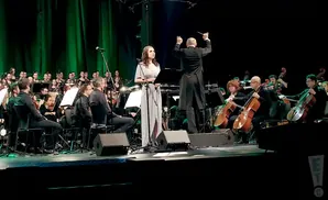 A female singer and conductor lead an orchestra and choir on a stage with green backdrops.