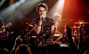 A male singer in a floral shirt performs on stage, holding a microphone.