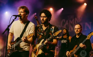 A band performs on stage with two guitarists in the foreground, a bassist in the middle, and a drummer in the background.