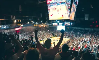 Fans cheer with fists raised in a packed basketball arena, watching the game on a large overhead screen.