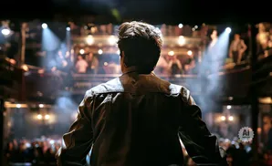 Man in denim jacket stands facing a blurred audience in a theater with spotlights.