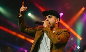 A man in a brown jacket and black cap sings into a microphone, pointing his finger upwards.