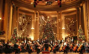 An orchestra performs in front of a large decorated Christmas tree and smaller trees in a grand hall.