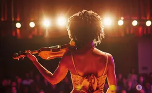 A woman plays the violin on stage, illuminated by spotlights, with an audience blurred in the background.
