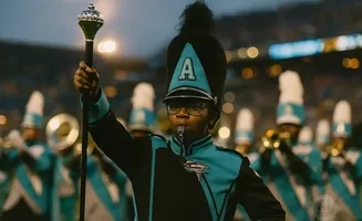 A young drum major in uniform leads a marching band, holding a mace and blowing a whistle.