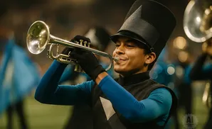 A band member in uniform plays a trumpet, wearing a tall black hat and blue sleeves.