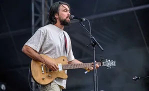 A man with a beard and long hair plays an electric guitar and sings into a microphone on stage.