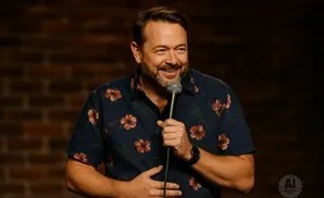 A smiling man with a beard in a floral shirt holds a microphone.