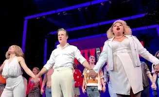 A group of actors on stage, holding hands, with the lead woman in a white dress and the lead man in a white shirt and pants.