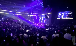 A concert crowd watches performers on a stage under bright purple lights, with screens showing live action.