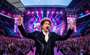Bob Dylan in a hat plays a piano on stage with another musician.