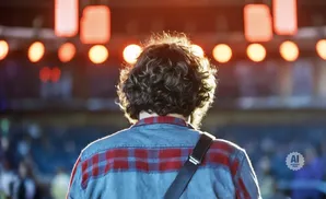 A person with curly hair wears a red and blue plaid shirt on stage with bright lights behind them.