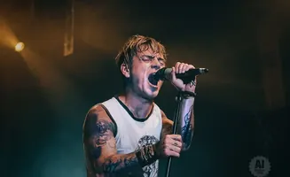 A tattooed man with blonde hair sings passionately into a microphone on a stage.