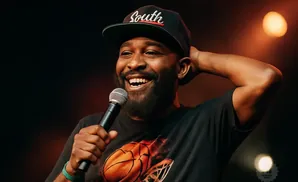 A Black man in a cap and t-shirt laughs while holding a microphone, with his hand behind his head.