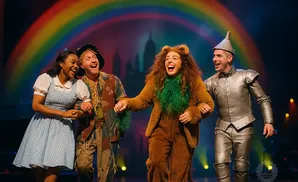 Dorothy, Scarecrow, Lion, and Tin Man from The Wizard of Oz laugh together on stage.