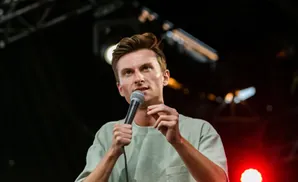 A man with light skin and brown hair holds a microphone and wears a light green t-shirt.