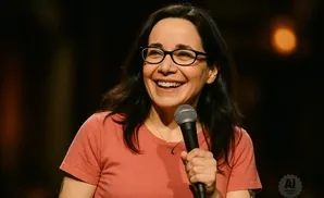 A woman with dark hair and glasses smiles while holding a microphone.