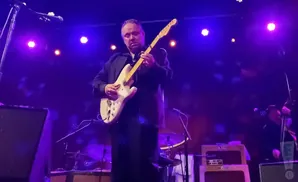 A man plays a white electric guitar on a stage with purple lights.