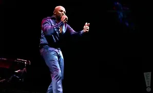 A bald Black man in a shiny blue shirt and pants sings into a microphone while gesturing with his free hand.