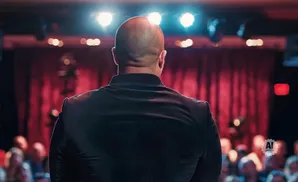 Man in a black suit facing a red curtain and bright stage lights with an audience in the foreground.