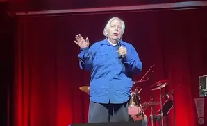 A man in a blue shirt speaks into a microphone on a red stage.