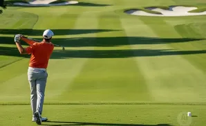 A golfer in an orange shirt swings at a golf ball on a sunny day.