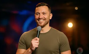 A man with short dark hair and a beard smiles while holding a microphone on a stage with blurred colorful lights.
