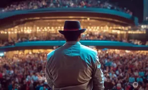 Man in fedora on stage facing a large, blurred audience in a theater.