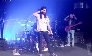 A male singer with long hair holds a microphone on stage, while another man plays a red bass guitar and a drummer is visible in the background.