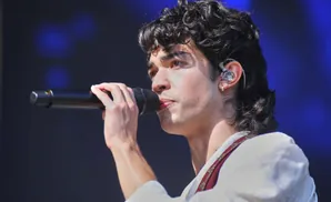 A male singer with curly dark hair holds a microphone to his lips while wearing an earpiece.