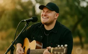 Man in black cap and jacket laughs while playing an acoustic guitar by a microphone outdoors.
