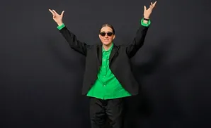 Person in a black suit and bright green shirt, arms raised, wearing sunglasses.
