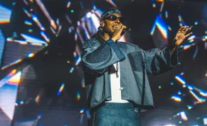 A man in sunglasses and a bandana performs on stage with a colorful, abstract background.
